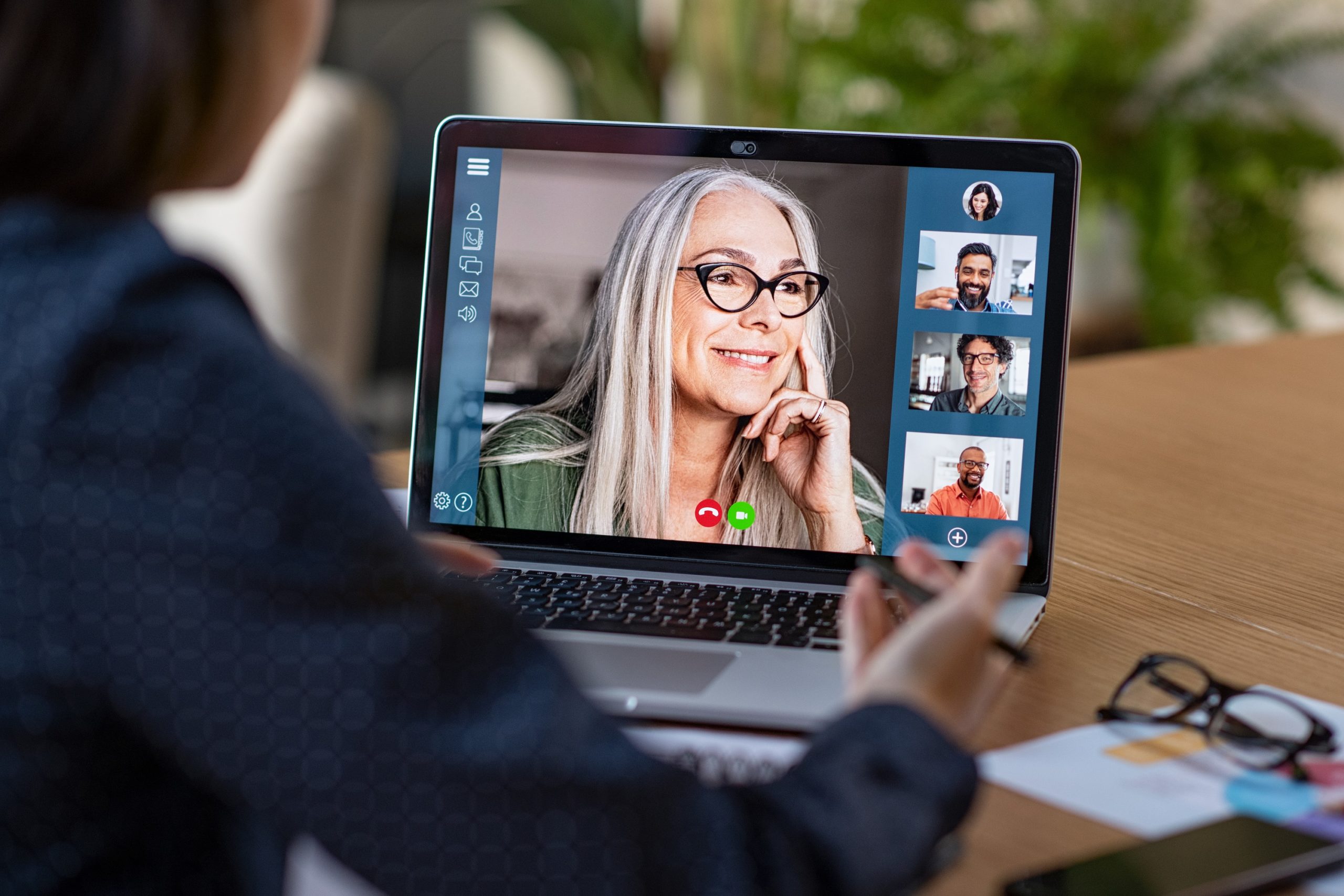 Multiethnic business team having discussion in video call. Rear view of business woman in video conference with boss and his colleagues during online meeting. Senior woman making video call with partners using laptop at home: remote job interview, consultation, human resources concept.